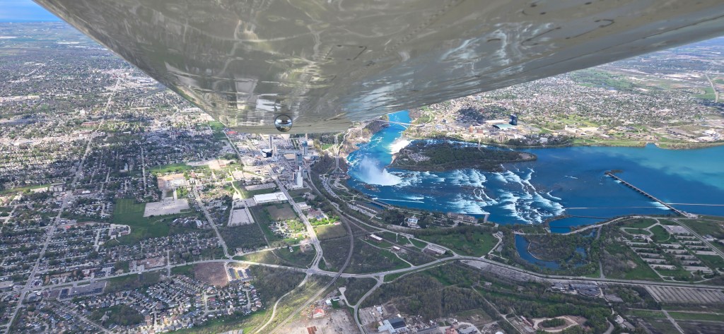 Aerial view of a city with a river and waterfalls, showing urban areas, greenery, and a blue sky.